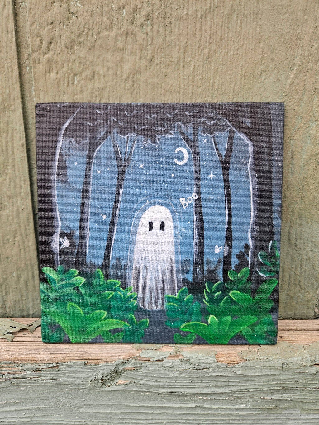 A small square canvas painting of a spooky forest at night with a white ghost, green bushes, stars, and a crescent moon, displayed against a rustic wooden wall.