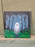 A small square canvas painting of a spooky forest at night with a white ghost, green bushes, stars, and a crescent moon, displayed against a rustic wooden wall.